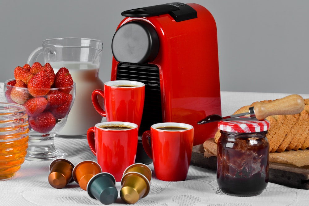 red coffee maker with single-use pods, red mugs, bread, strawberries and jam on white surface