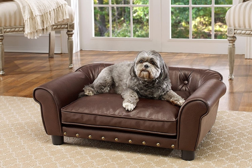 A small dog is sitting on a dog bed that looks like a mini brown leather couch.