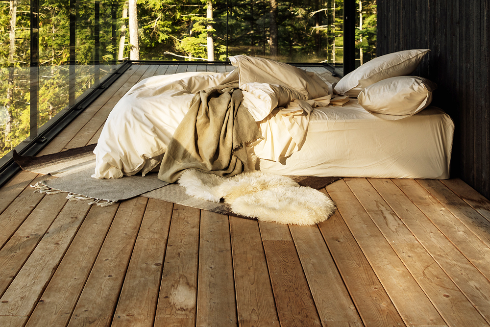 comfy bed on wood floor in front of glass railing with green trees and mountains surrounding