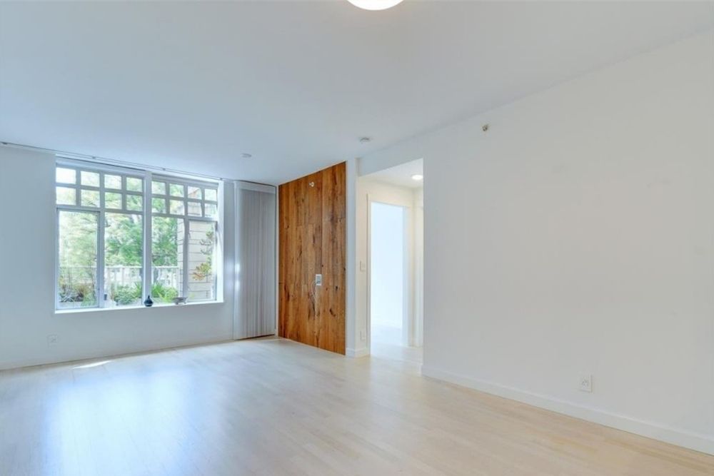 An empty condo main living space with white walls, a window, a wood accent feature and light hardwood floors.