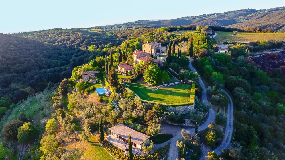 An expansive medieval castle atop rolling green hills in Italy with a view of the moutains