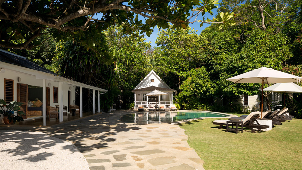 White villa surrounded by patio furniture, small pool and trees providing shade