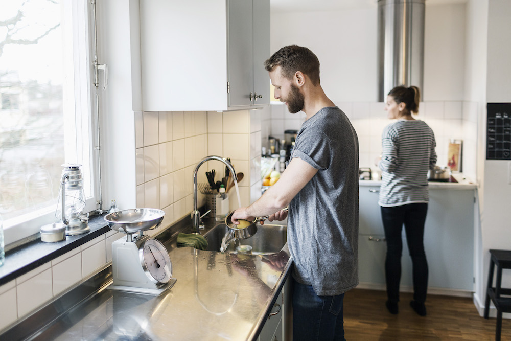 man washing sauce pan while woman standing in background in modern white kitchen