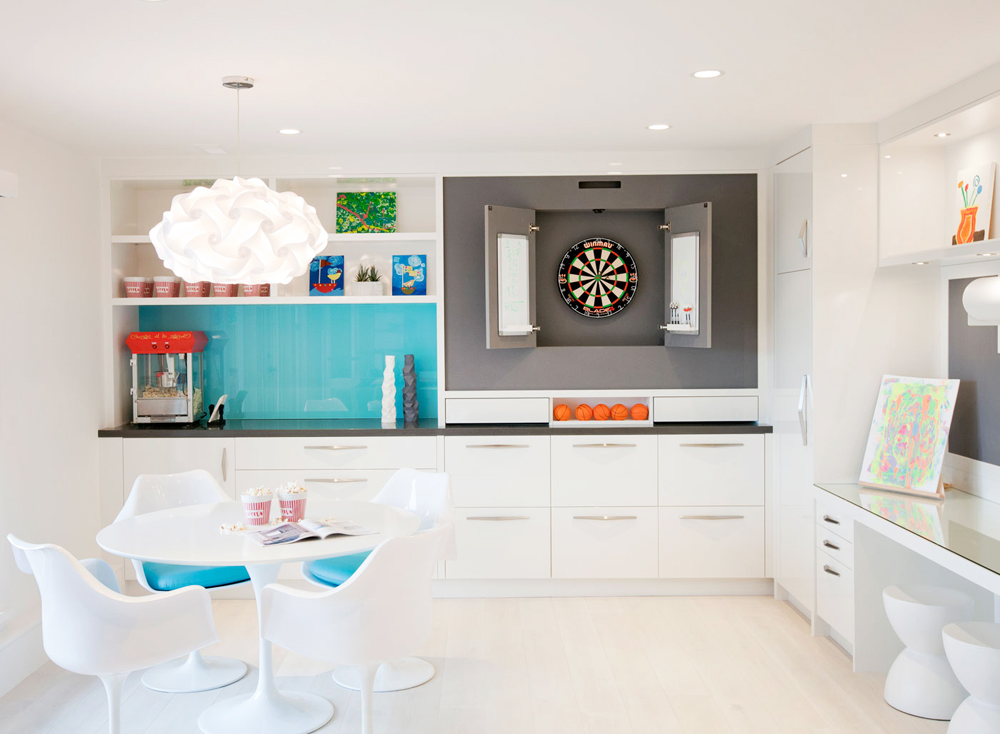 A white renovated basement game area with a dart board and popcorn machine with pops of colour scattered throughout