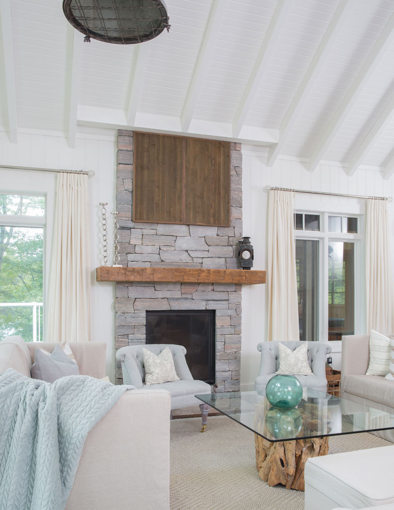white living room with stone front fireplace with wood mantel and wood cupboards flanked by curtained windows