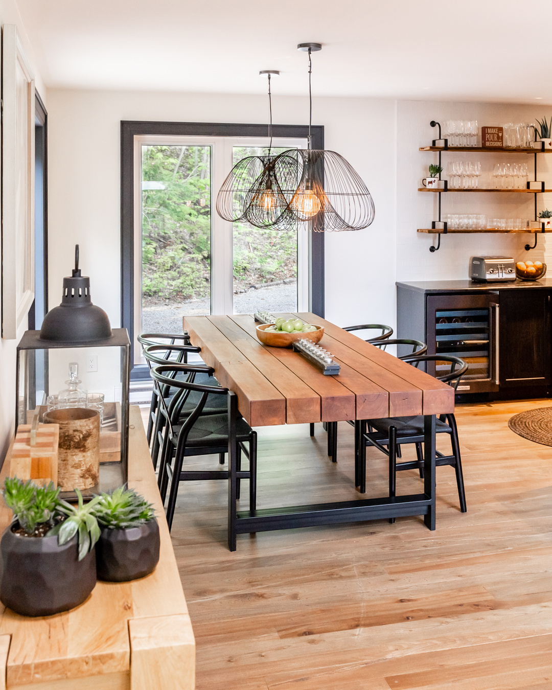 wood topped dining table with black legs and chairs, black wire pendant lights, kitchen with hanging shelves to the right