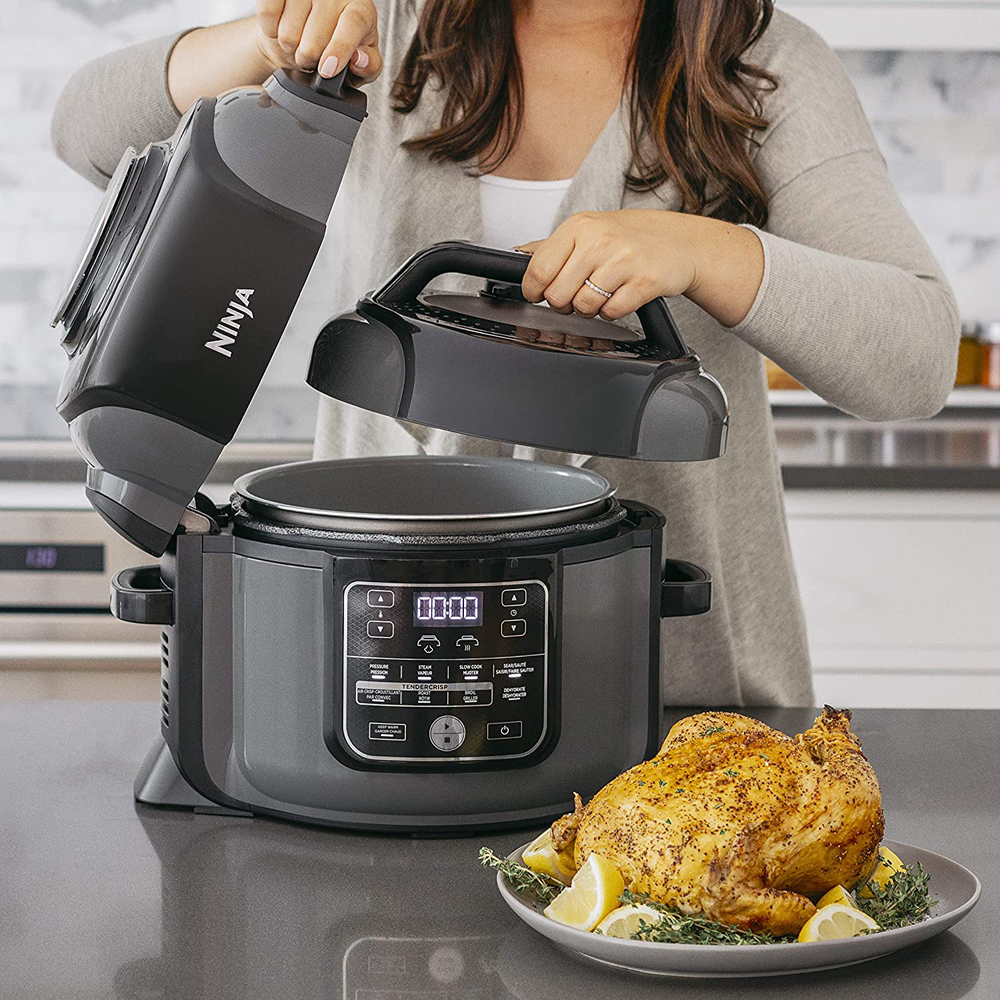 A woman opens the lid of her grey Ninja pressure cooker in preparation of putting a whole seasoned chicken inside