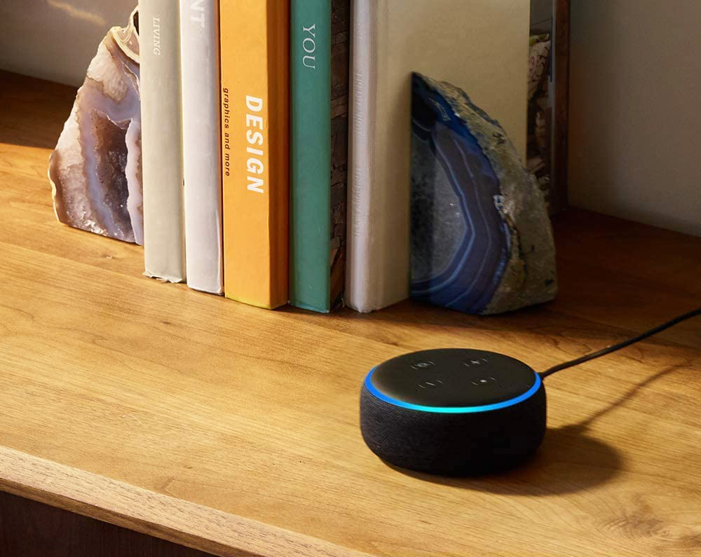 A fabric design black smart speaker with Alexa sitting on a wooden cabinet next to a stack of books