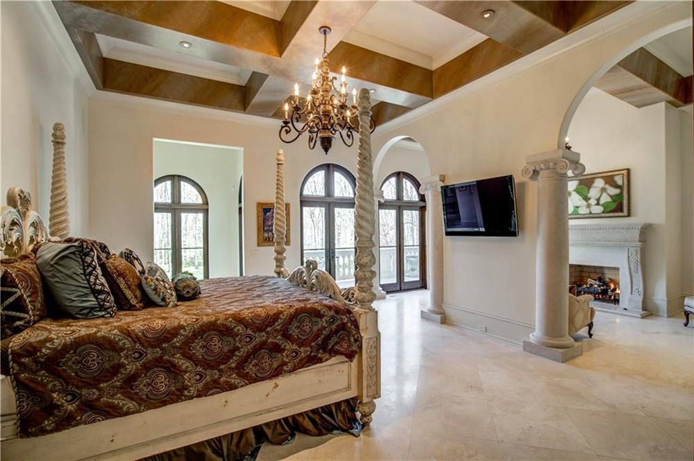 A spacious master bedroom with ornate bed columns, low-hanging chandelier and a separate seating area with a wood-burning fireplace