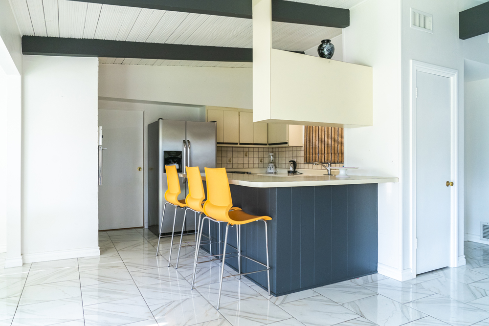 A tiny, outdated kitchen nook with old tiles, eye-level cabinets, yellow chairs and navy blue island