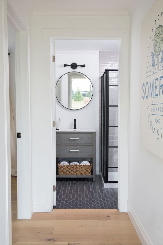 view into white, black and grey bathroom with round mirror reflecting a pine tree