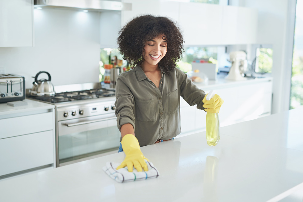 A Black woman wearing yellow rubber gloves in a clean white kitchen, wiping down the island counter