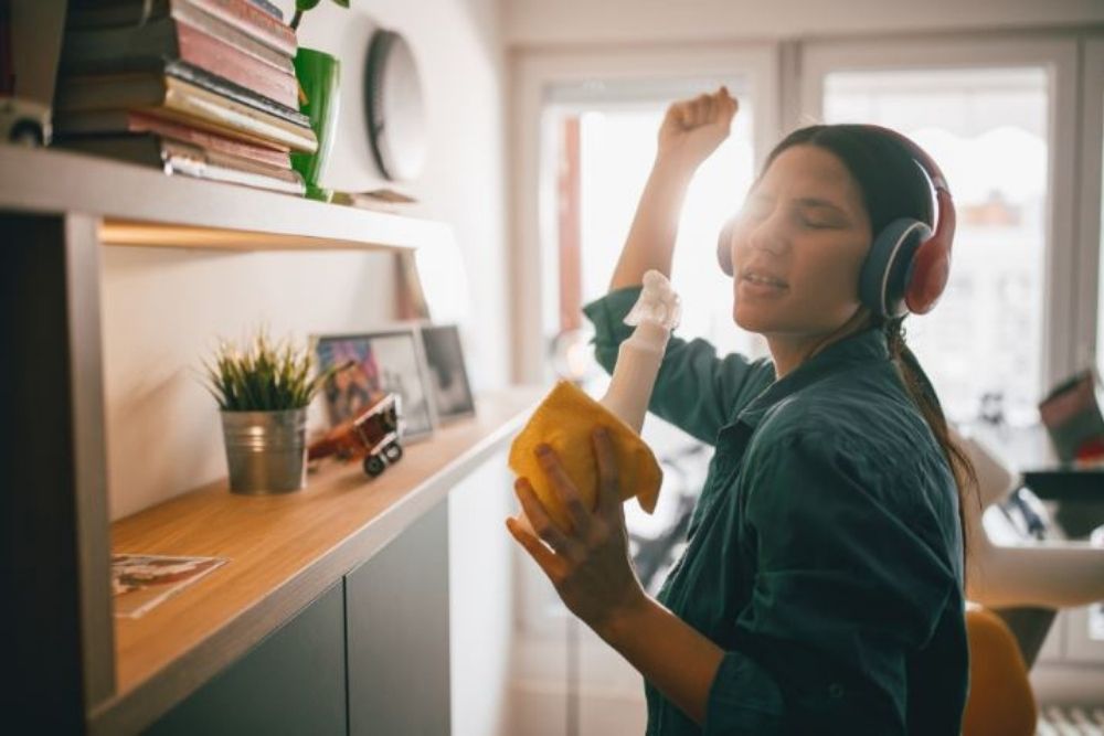 A young Asian woman wearing headphones and dancing around her apartment as she cleans