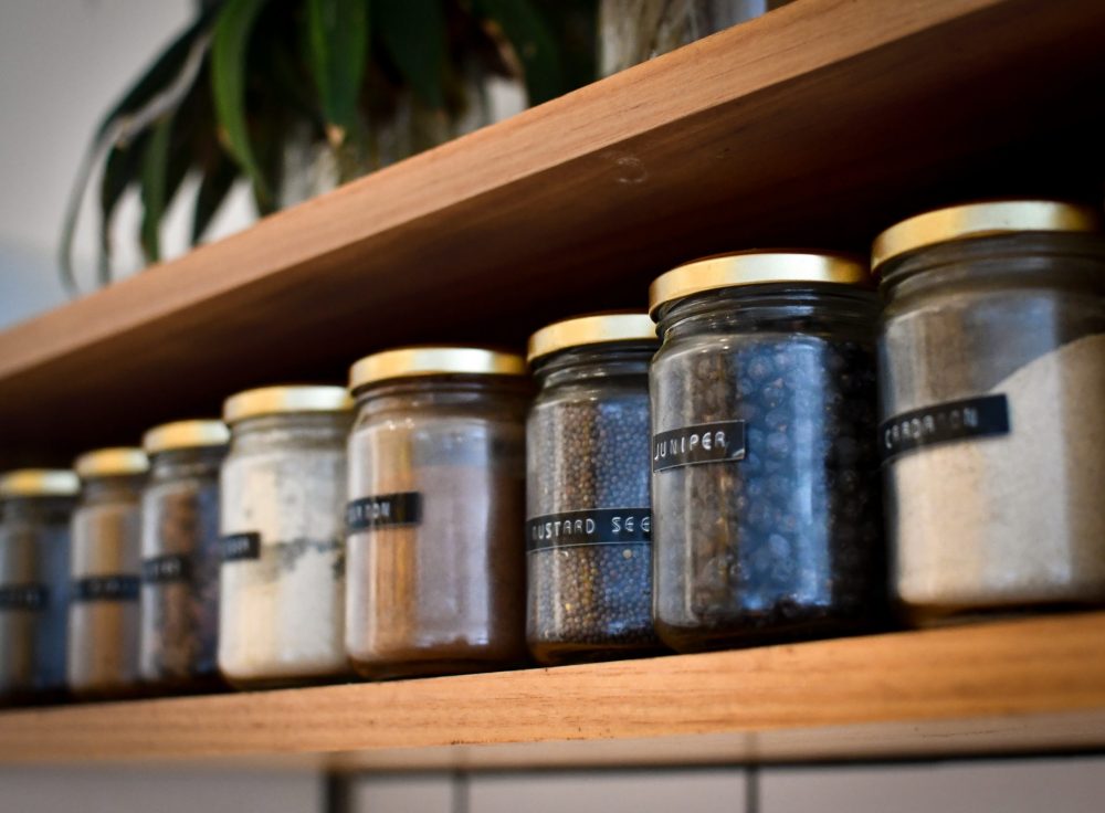 A row of glass jars on a wooden shelf in a kitchen, each clearly labeled