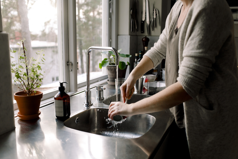 A white woman wearing a cardigan, washing a mug in the sink that is situated in front of a window