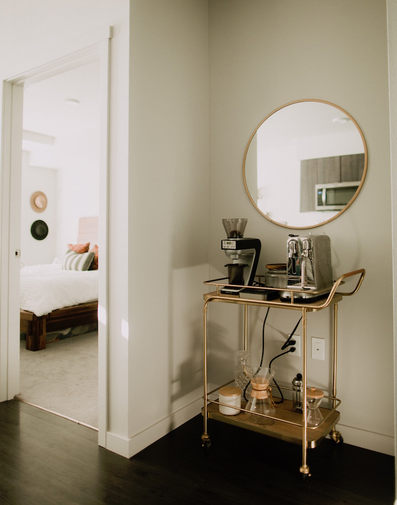 Metallic bar cart with coffee machines and supplies in front of round mirror in corner