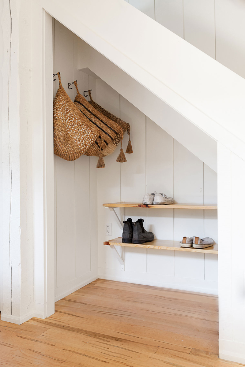 A white nook under the stairs of a house with shelves for shoes and hooks for hanging bags and jackets