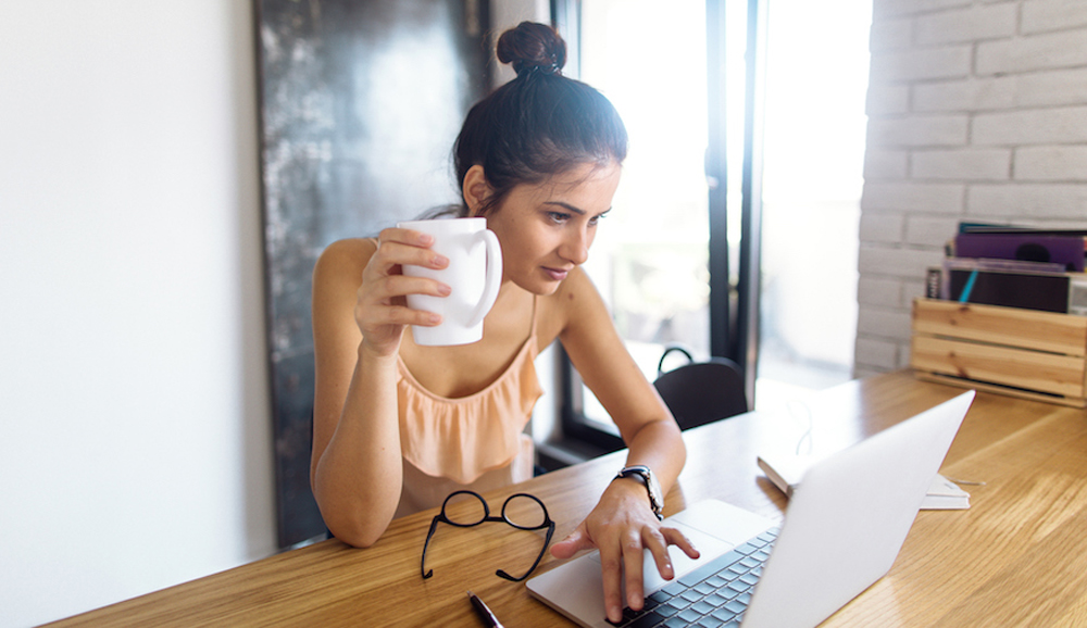 A young woman holding a mug of coffee while sitting at the kitchen table with her laptop