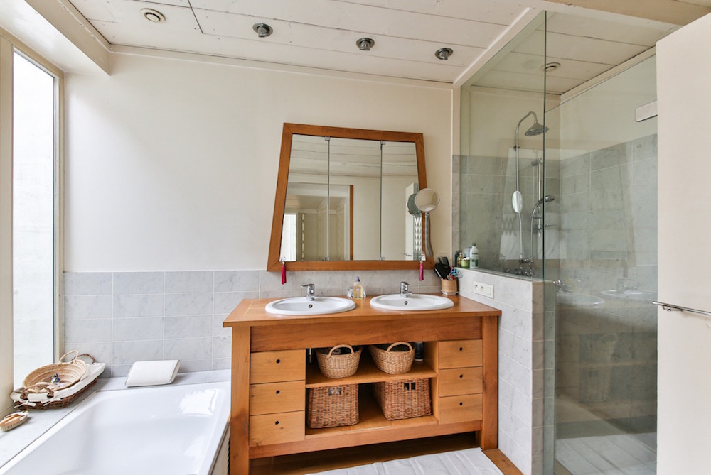 A small, narrow bathroom with a wood vanity, storage space, standing shower and separate soaker tub next to the window