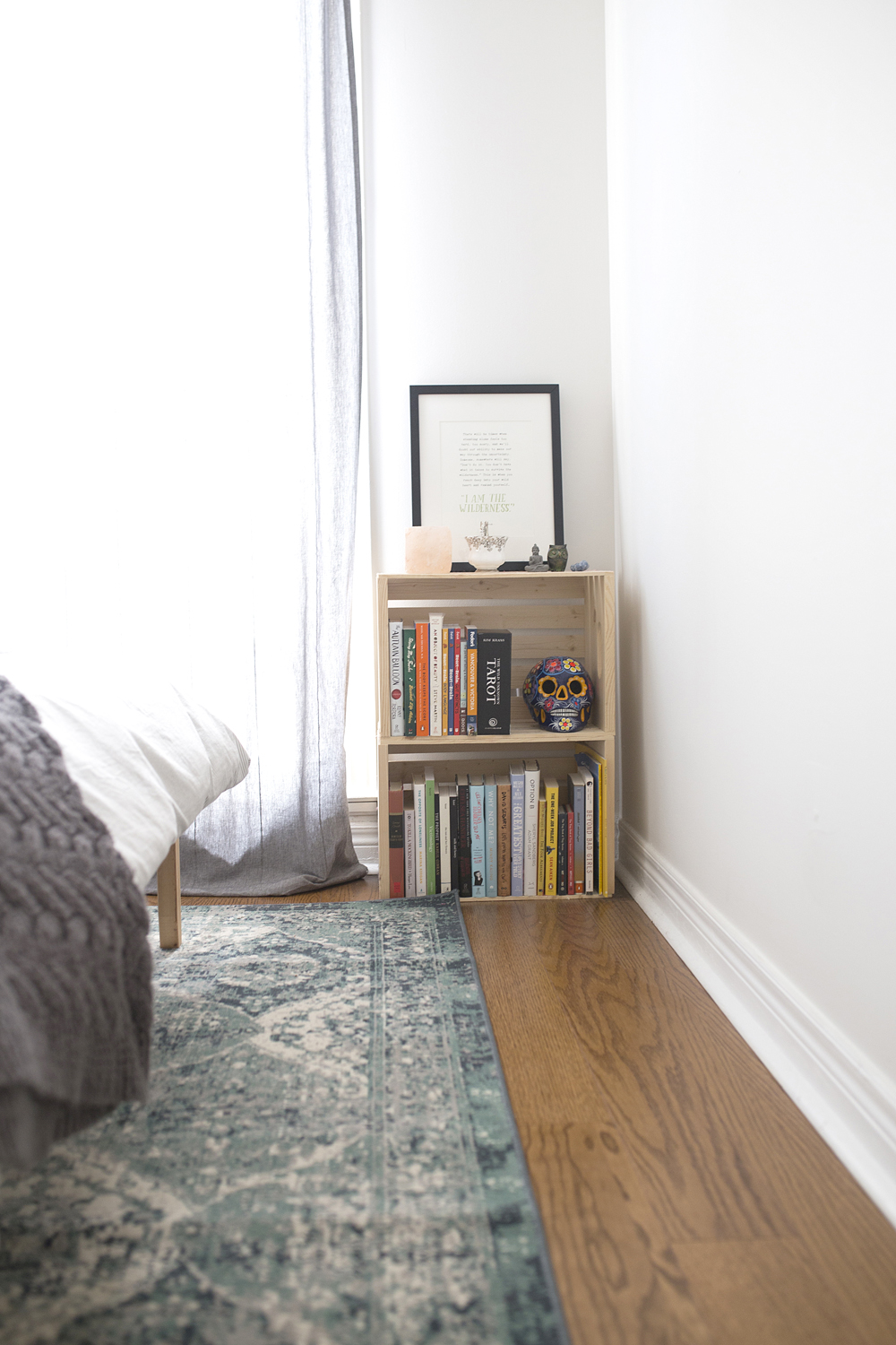 A sunny bedroom with a small crate bookshelf on the floor and filled with books and knick-knacks