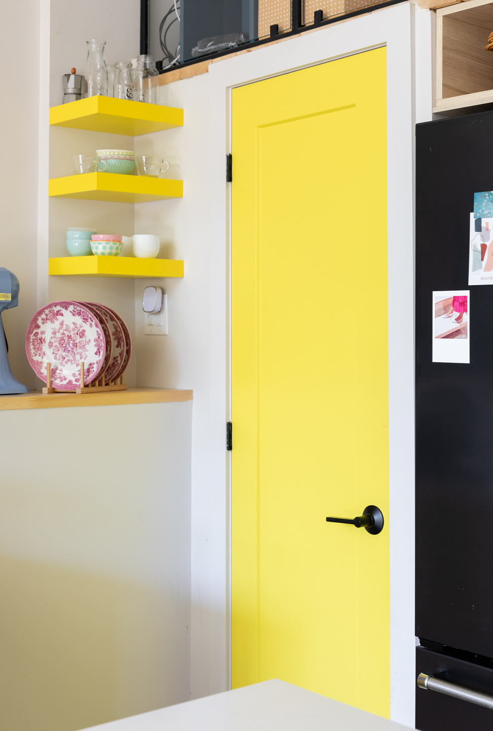 A bright lemon-yellow door in a white room next to yellow open shelves
