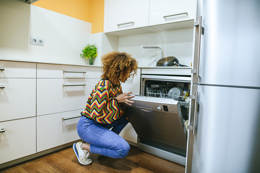 A young woman kneels in front of her dishwasher
