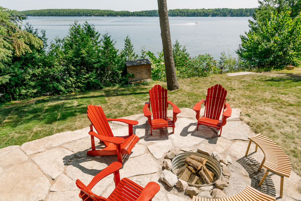 A rustic backyard fire pit near the lake and surrounded by Muskoka chairs
