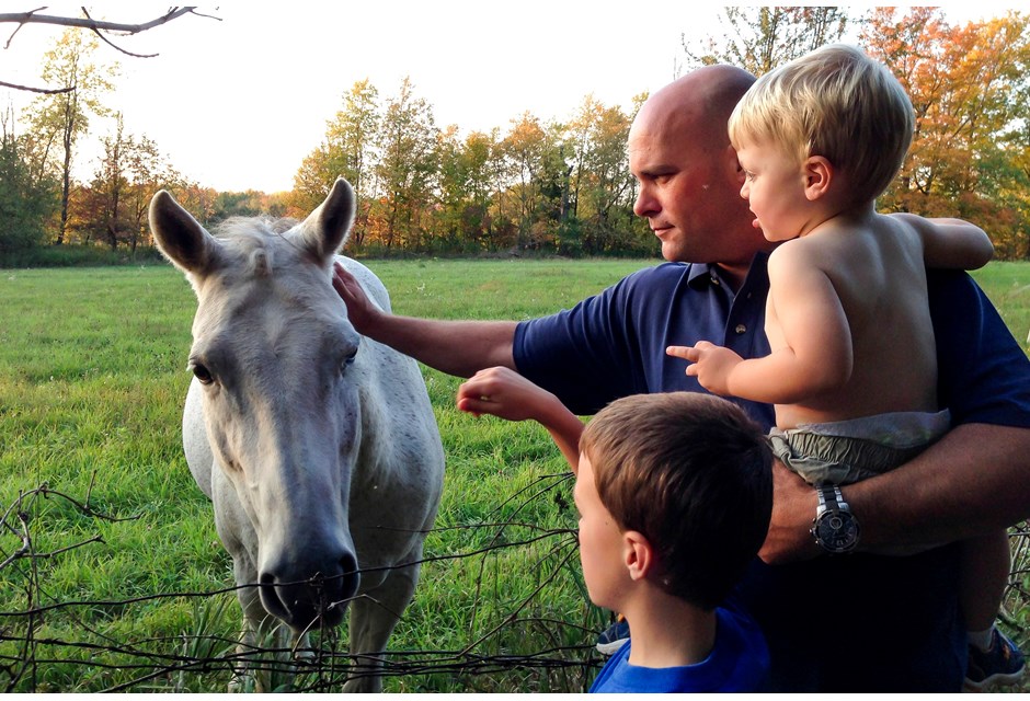 Bryan Baeumler, and his sons Quintyn and Lincoln petting a white horse, in an open green field.