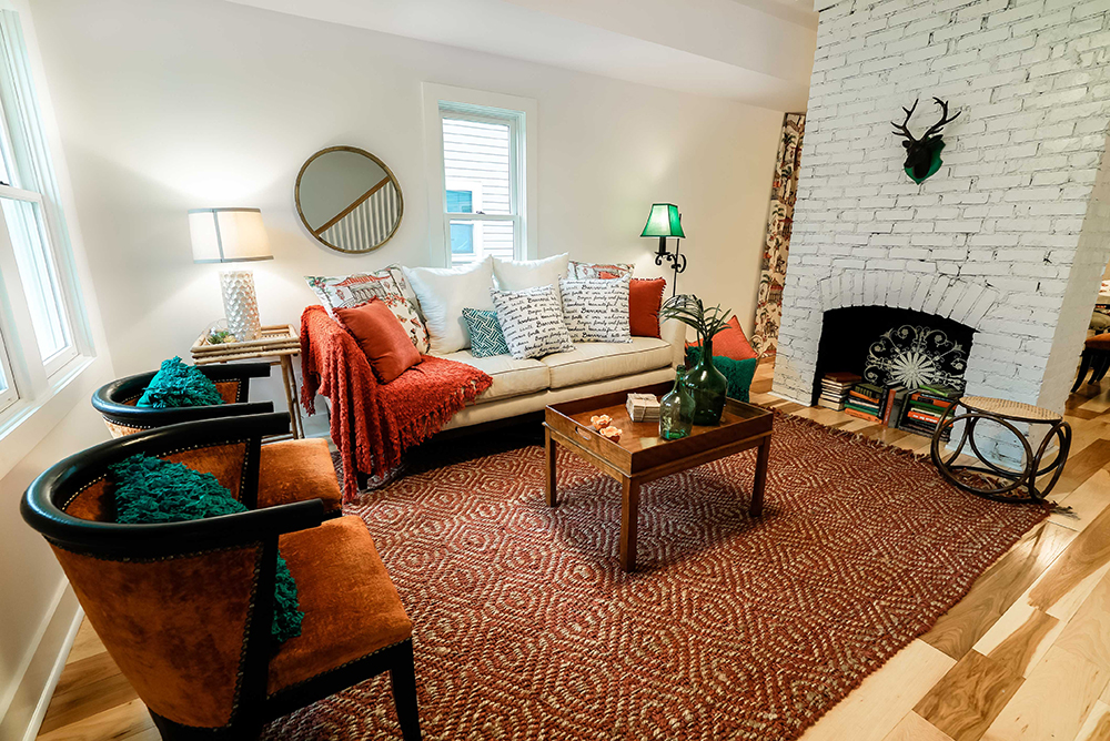 Modern living room with geometric rug and warm-toned furniture. Inside a whitewashed brick fireplace is a stack of books.