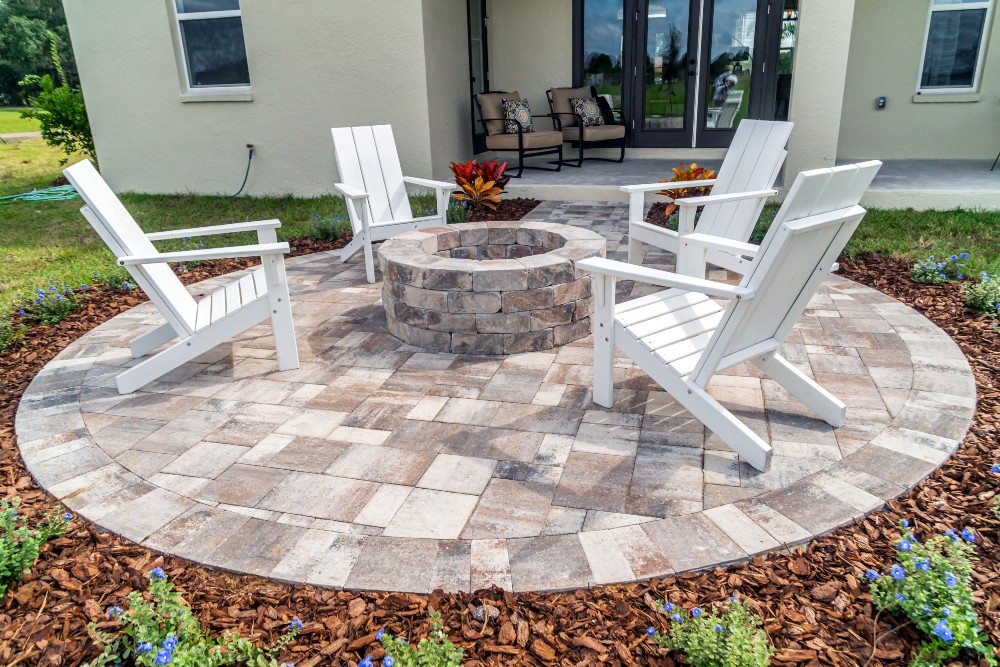 A stone patio and firepit area with four white Adirondack chairs.