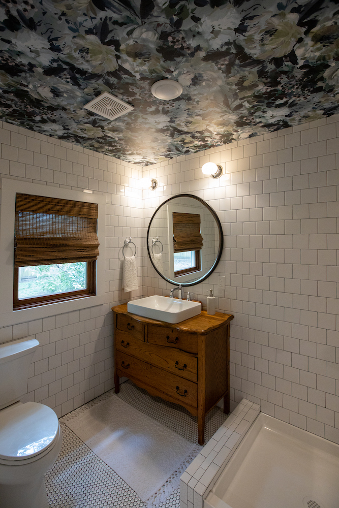 white-tiled bathroom with floral wallpaper on ceiling and wooden dresser vanity