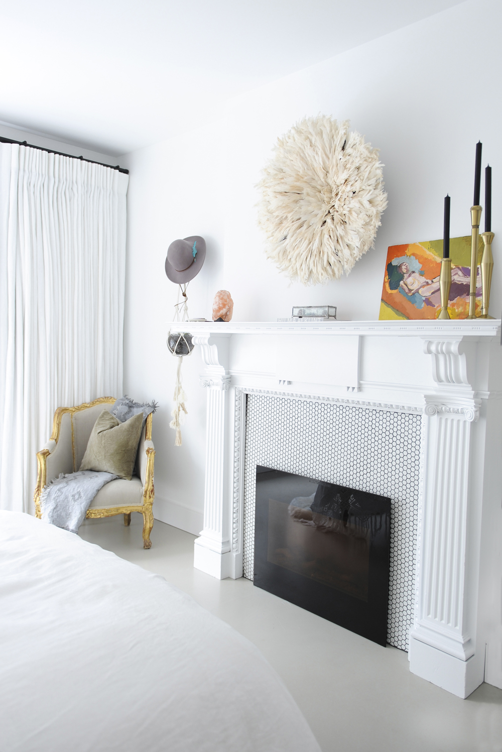 white bedroom with fireplace with mosaic tiles and feather juju above it, gold frame chair in corner