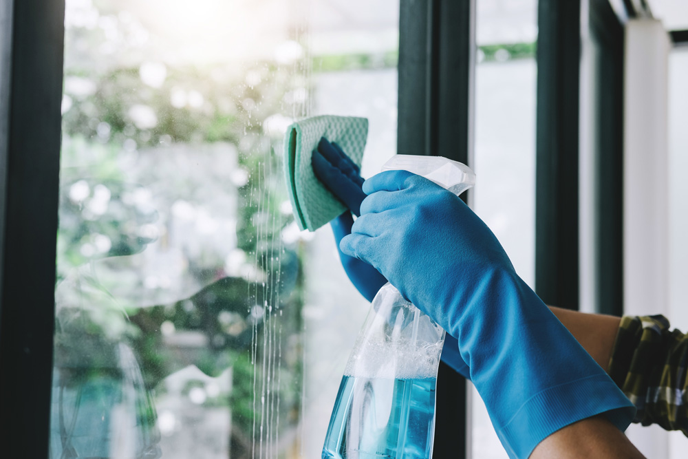 A blue rubber gloved hand cleans a large window with cloth and solution