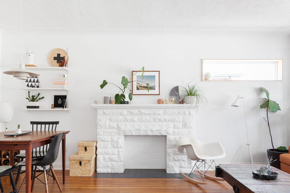 A clean white living room area with painted stone fireplace and various floating shelves, potted plants and artwork