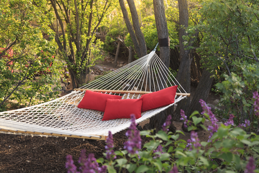 Spacious white hammock with bright red pillows strung between two trees in a forested backyard