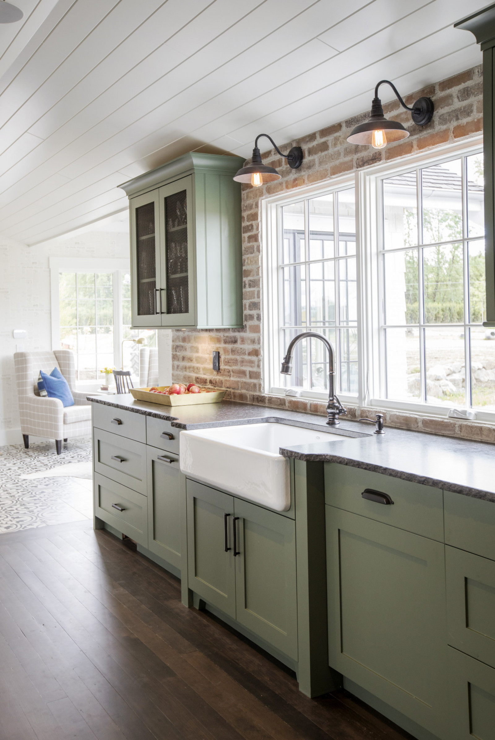 white apron sink in front of kitchen window with green cabinets and two sconces above