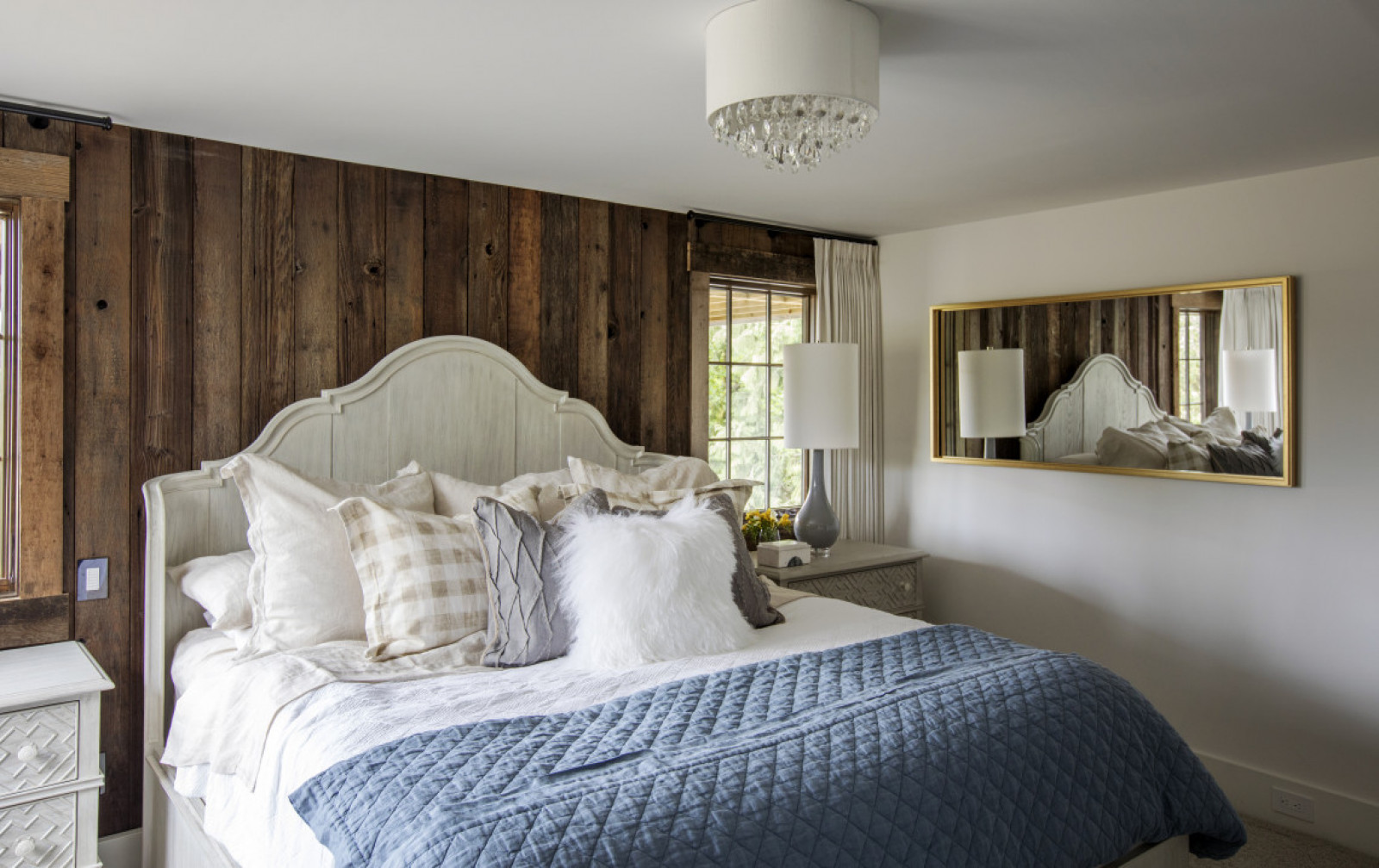 bedroom with wood plank wall, gold frame mirror on one wall, blue quilt, lots of cushions, white furry one in front