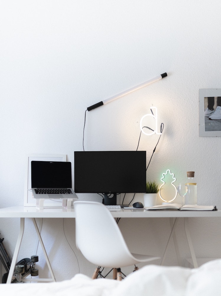 white desk in white home office with lots of electronics on top and small neon light on wall