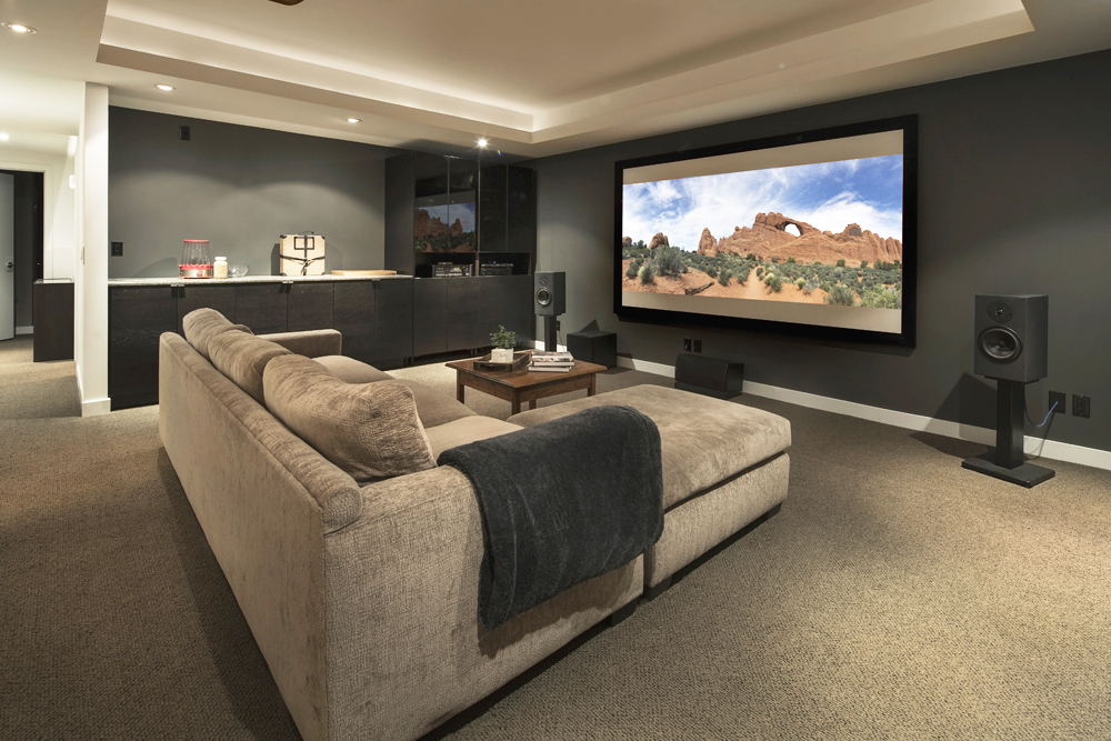A basement home theatre with a large screen, L-shaped couch and bar table off to the side