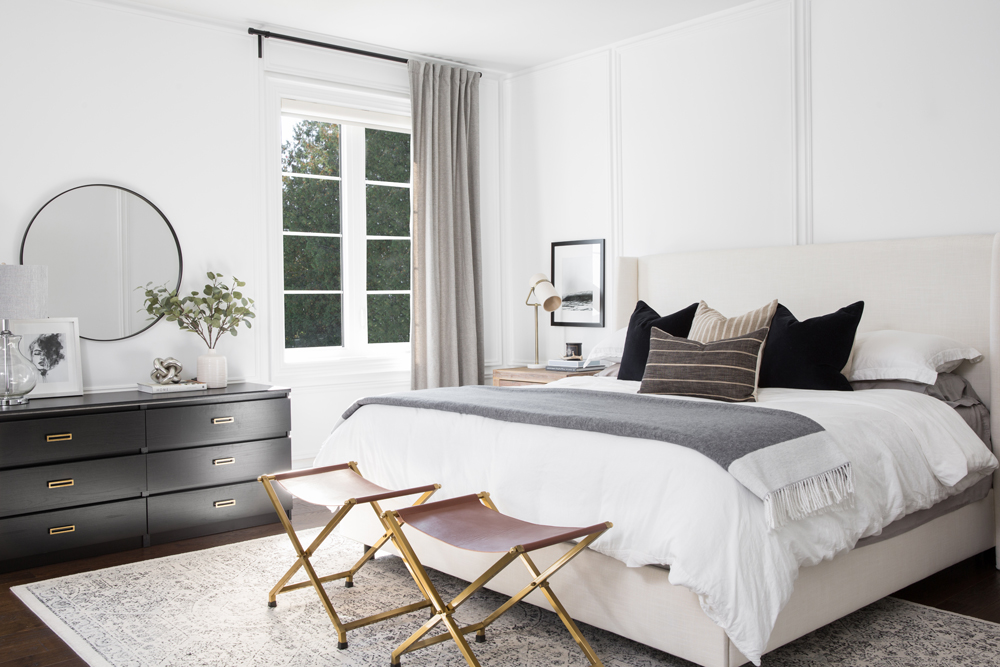 white bedroom, window with grey drapes, two brass and leather stools at bottom of bed