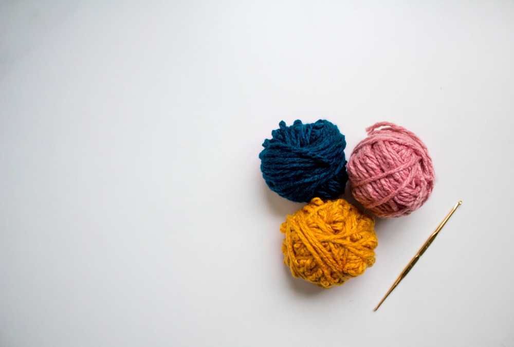 Three balls of yarn in blue, pink and mustard yellow next to a crochet needle against a while background