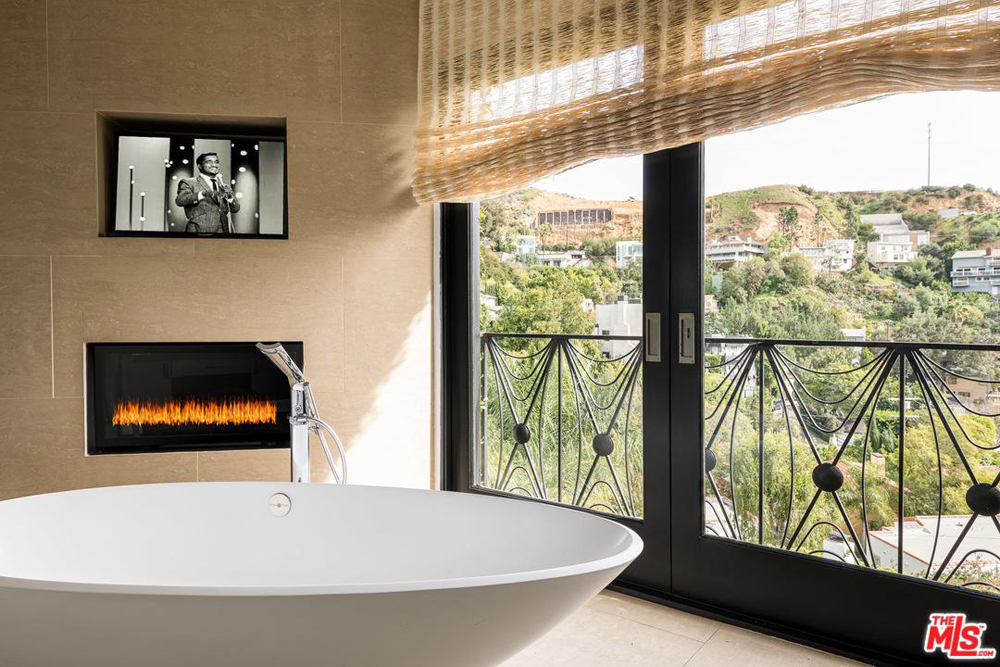 A private master bathroom with deep soaker tub, gas fireplace with a TV above it and Juliet balcony overlooking the Hollywood Hills