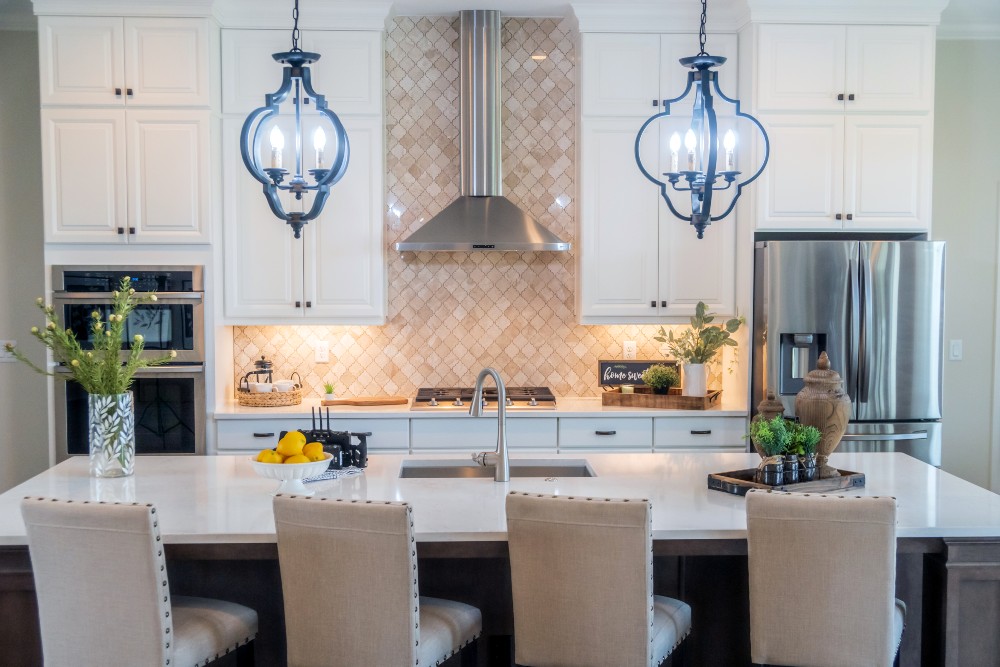 A modern kitchen with white cabinets, a shiny Arabesque tile backsplash, white countertops, stainless steel appliances, and an island with sink and upholstered stools