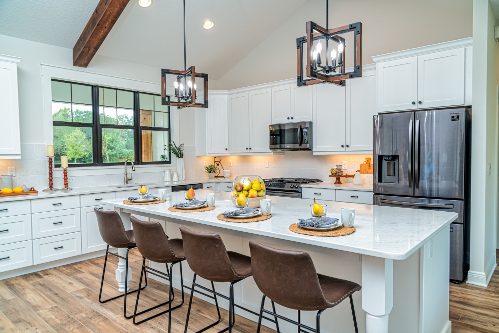 Farmhouse kitchen with white shaker cabinetry, white countertops, a long, rectangular islands with brown seating, stainless steel appliances, and wood-accented lighting fixtures.