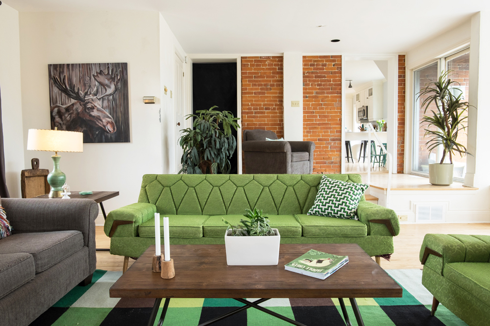 An avocado-green sofa in the centre of a minimalist living room with an exposed brick wall and plenty of indoor plants
