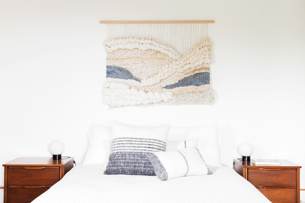 A handmade blue, grey and beige macrame wall piece hangs above a minimalist bed