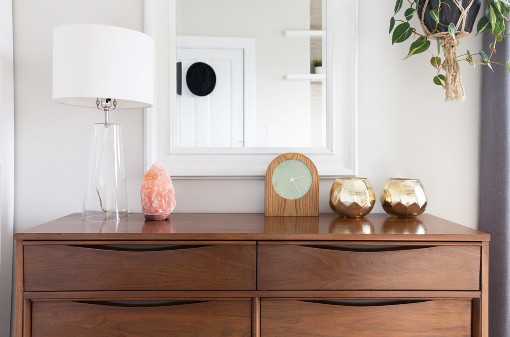 A pink Himalayan salt lamp sits on top of a bedroom dresser next to a clock and brass decorative vases