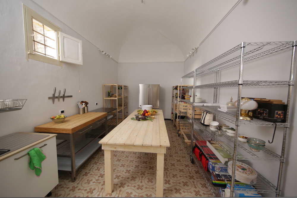 white prep kitchen with metal shelving units, large stainless steel fridge and wooden table
