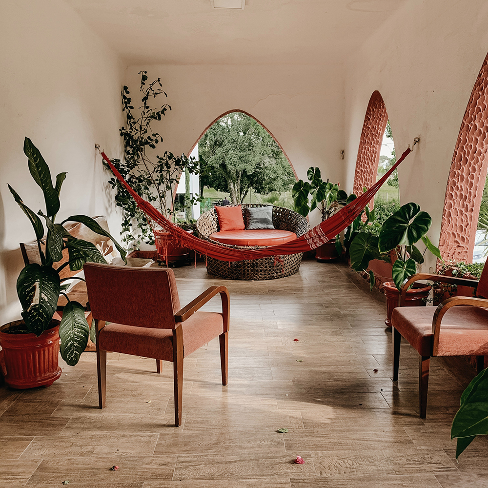 Covered porch with large potted plants, boho furniture, and a red hammock in the middle of the room.