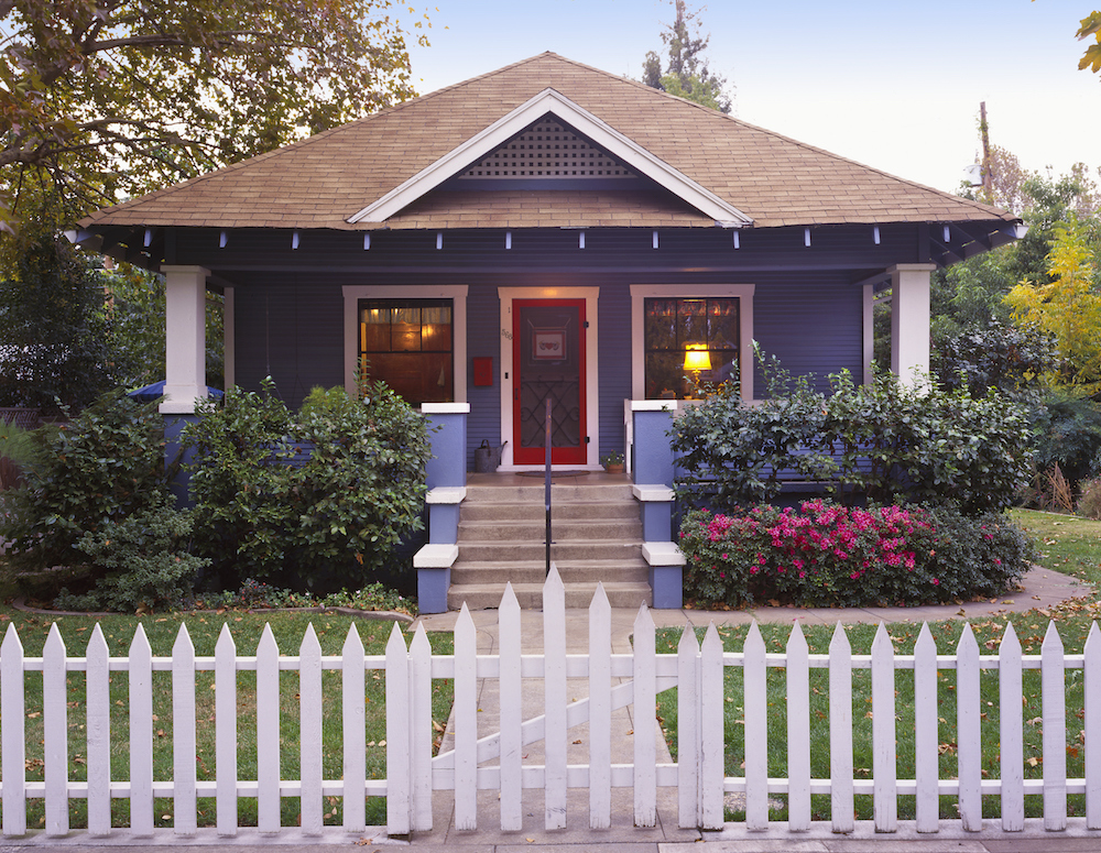 A small blue craftsman bungalow with a white picket fence and surrounded by flower bushes